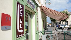 A hotel entrance with a green awning, a signboard with the hotel's name, and a street view with parked cars and a pedestrian. - Le Lion D'or (Gémozac)
