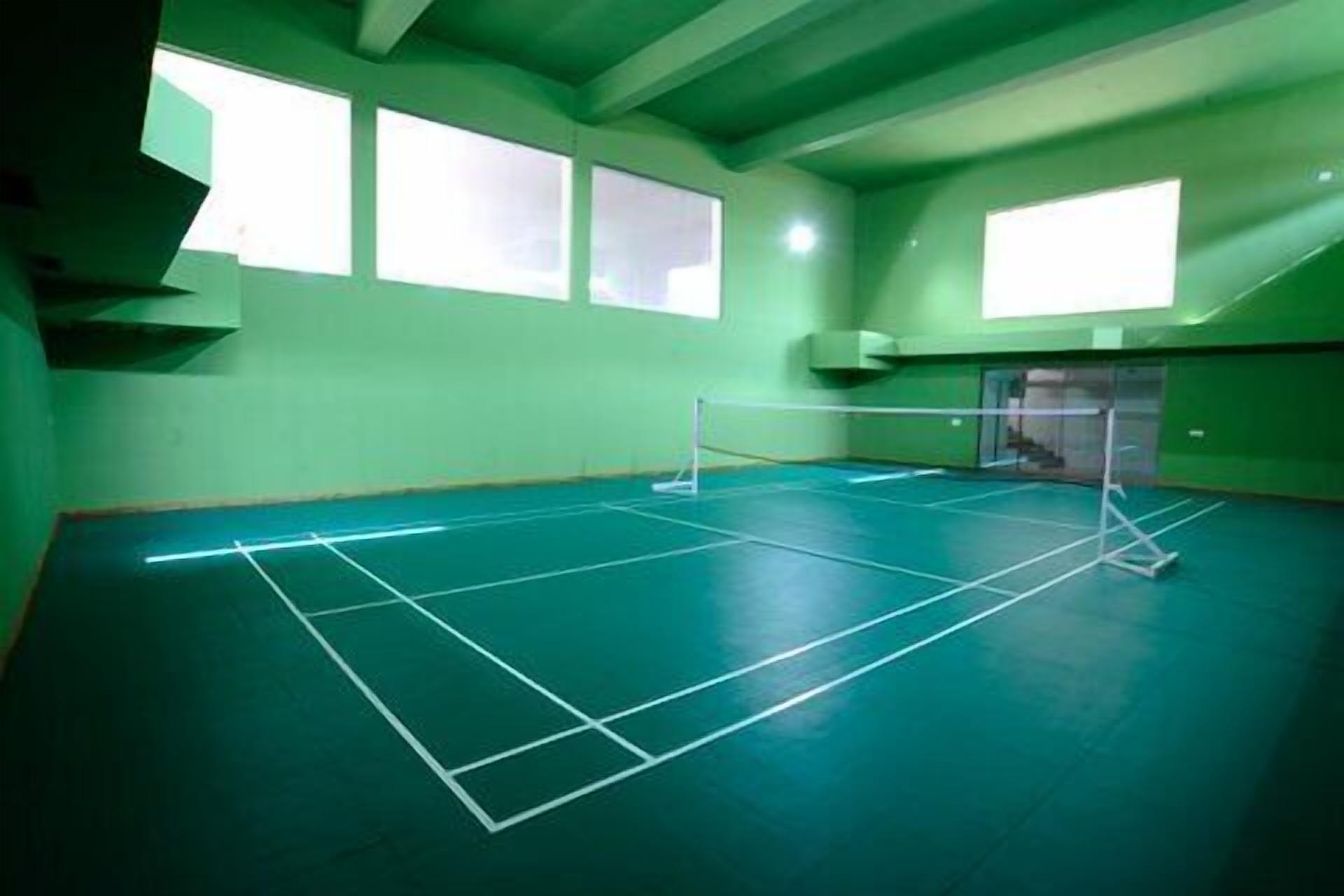 A tennis court with a net, green walls, and large windows.
