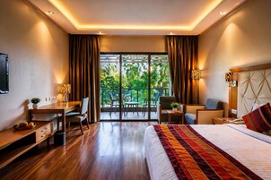 A hotel room with a bed, desk, chair, and a balcony with a view of greenery.