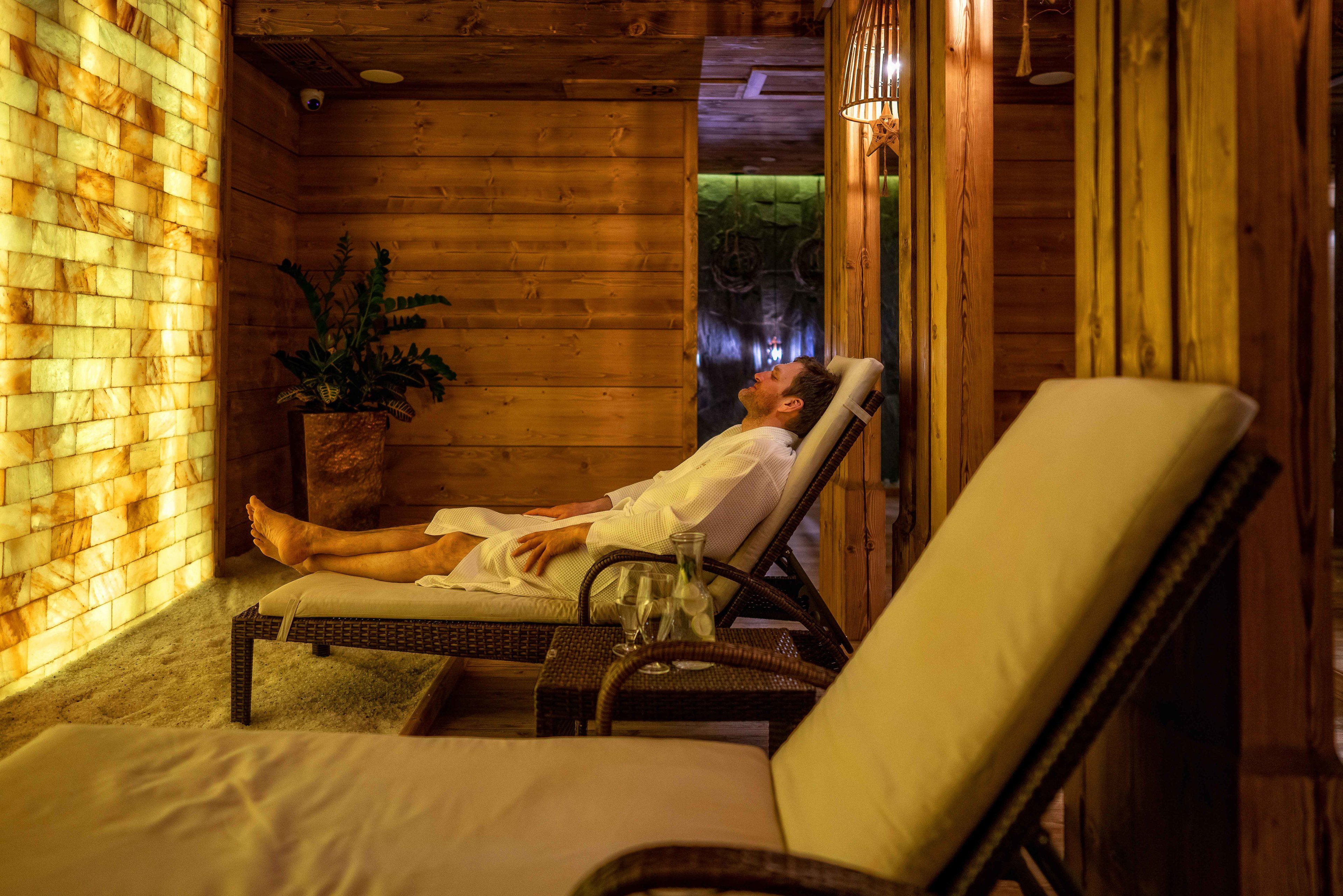A wooden cabin interior with a person relaxing on a wicker chair by a window.
