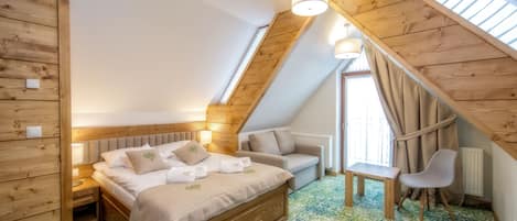Un dormitorio acogedor en el ático, con techo inclinado de madera, una cama con cabecera, una mesita pequeña, una silla y una ventana con cortinas.
