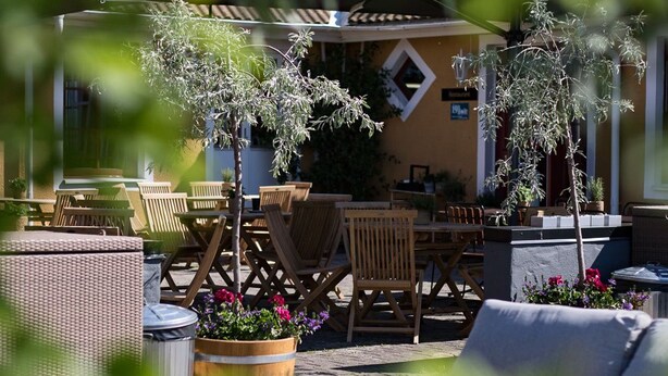 An outdoor seating area with wooden tables and chairs, surrounded by potted plants and a stone wall.