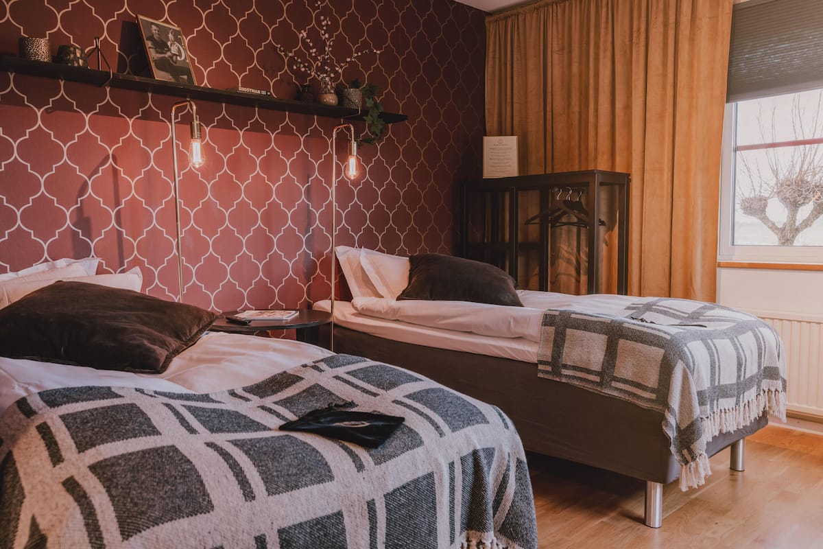 Een slaapkamer met twee bedden, een behang met patroon, een plank met decoratieve objecten en een raam met gordijnen.