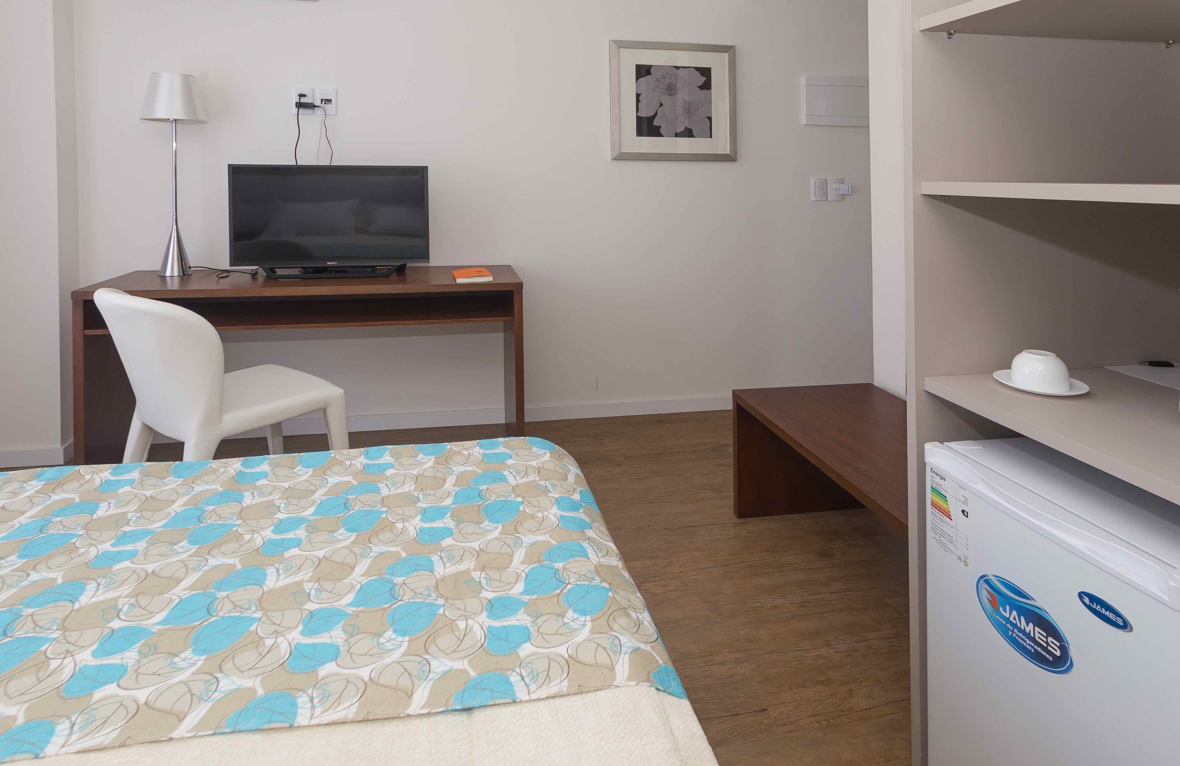 A hotel room with a bed, a desk with a television, a chair, a white refrigerator, and a picture on the wall.