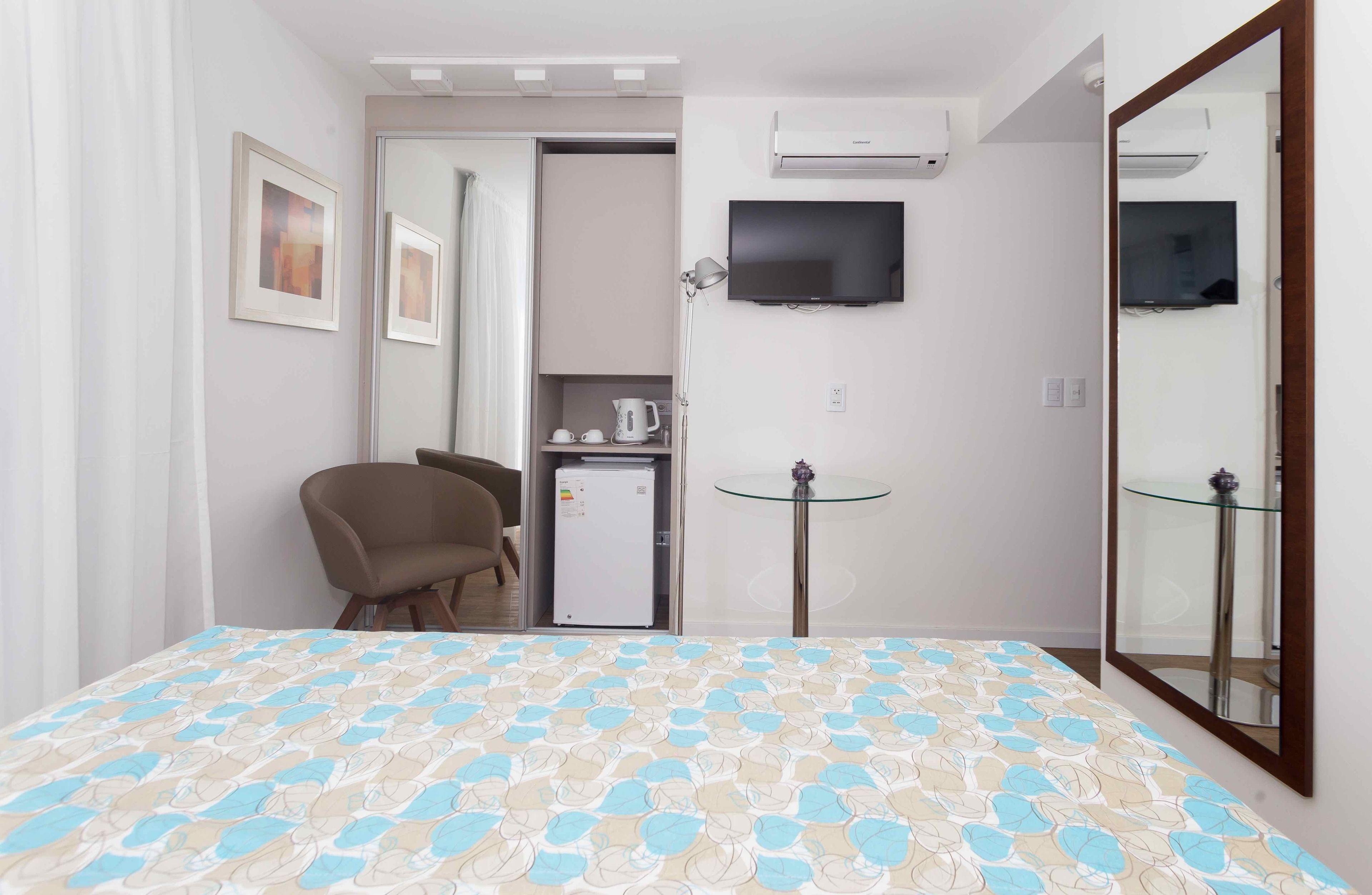A hotel room with a bed, a desk, a chair, a refrigerator, a television, and a mirror.