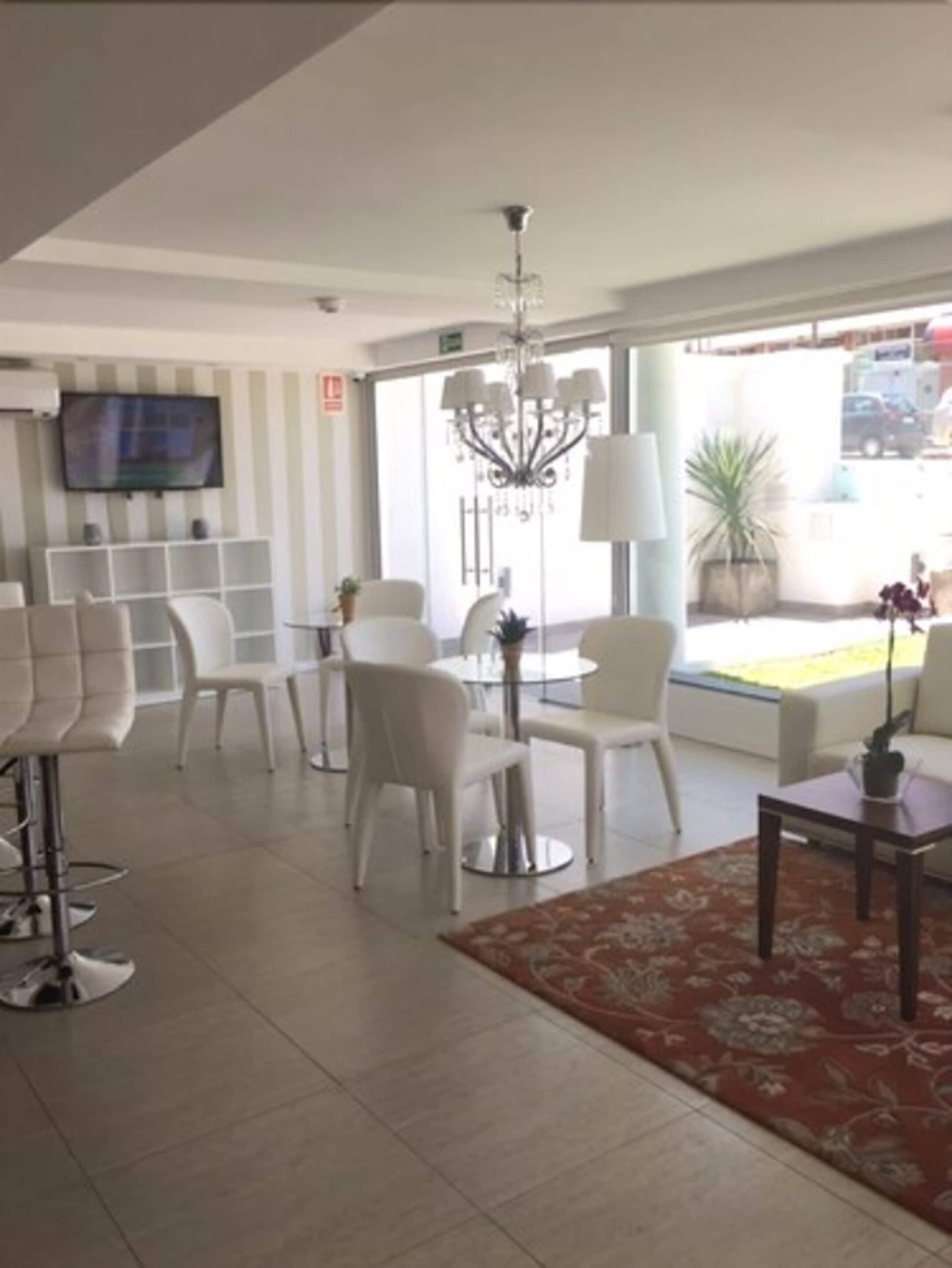 A modern hotel lobby with white furniture, a chandelier, and a TV mounted on the wall.