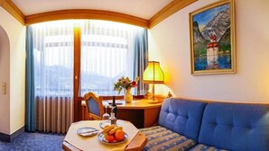 Une chambre d’hôtel avec une table à manger dressée, comprenant des assiettes, un panier de fruits et une lampe. On y trouve également un canapé et un tableau accroché au mur.