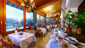 Un restaurant avec des tables dressées pour le service, de grandes fenêtres offrant une vue sur les montagnes et des plantes en pot.