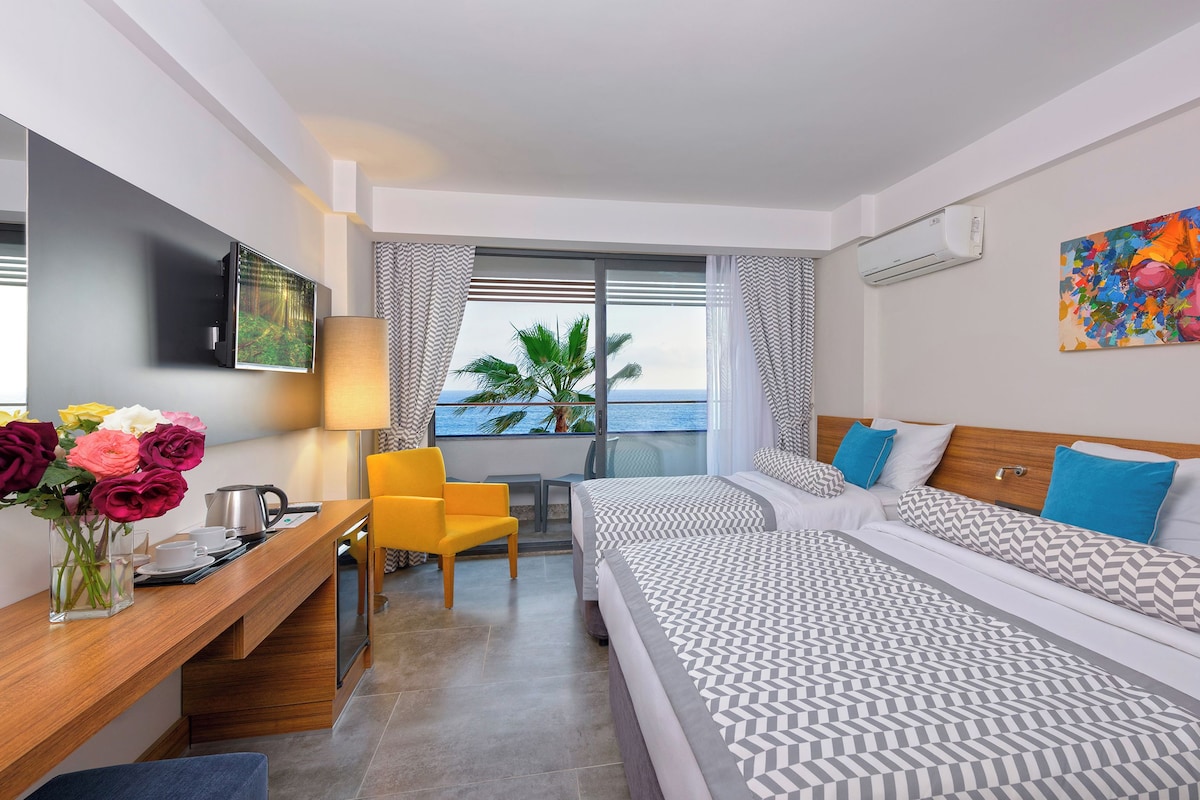 A hotel room with a bed, a desk, a chair, a TV, a painting, and a view of the sea.