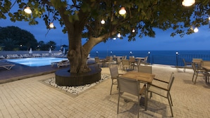 Zona de piscina exterior con sillones y mesas, rodeada de un árbol y vistas al mar.