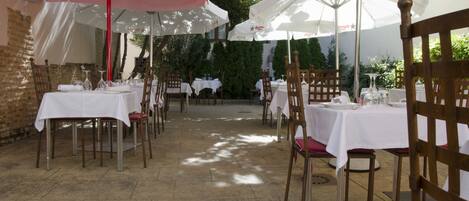 Un área de comedor al aire libre con mesas y sillas, cubierta por sombrillas, y un muro de ladrillo al fondo.