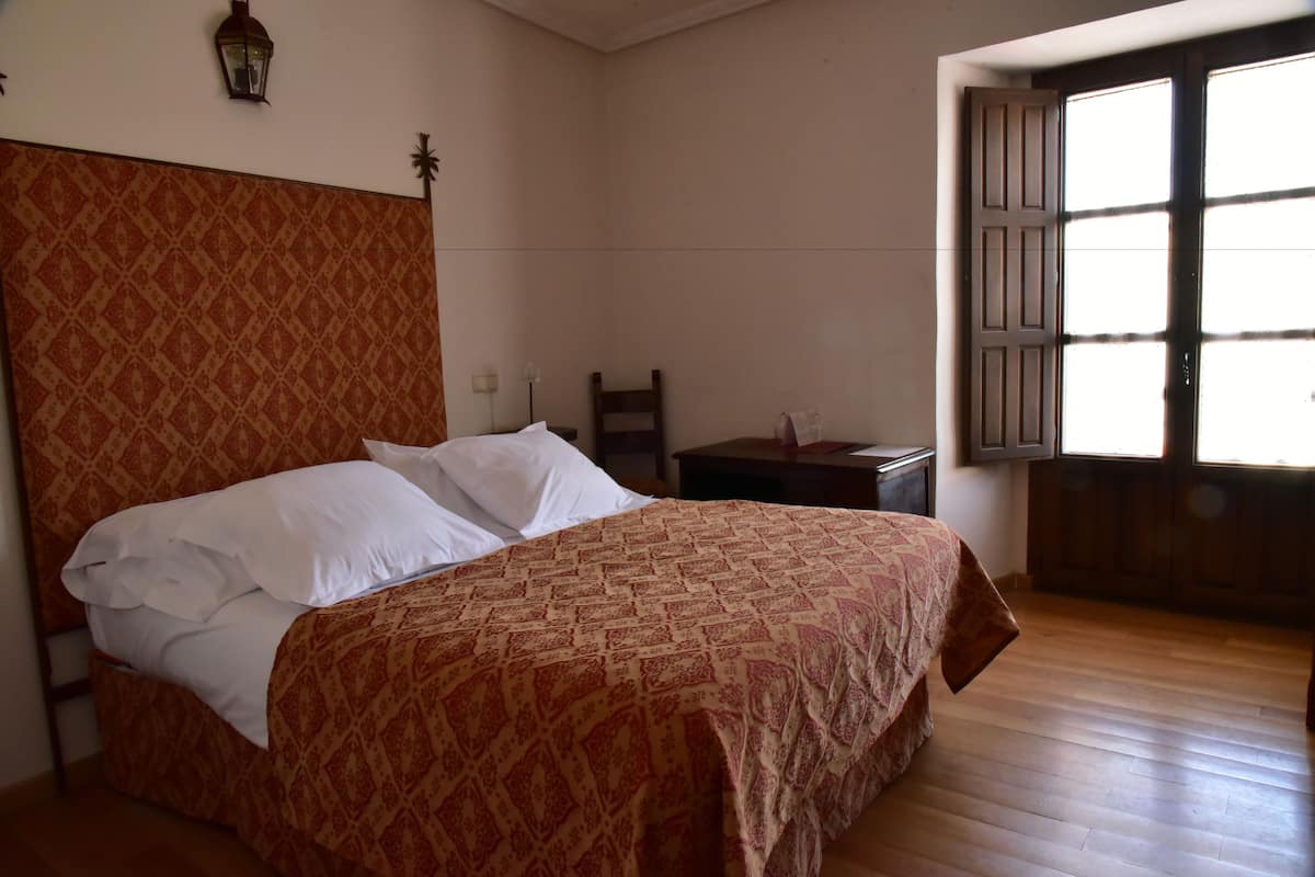 Un dormitorio con una colcha estampada, cabecera de madera, mesita de noche, una silla y una ventana con contraventanas de madera.
