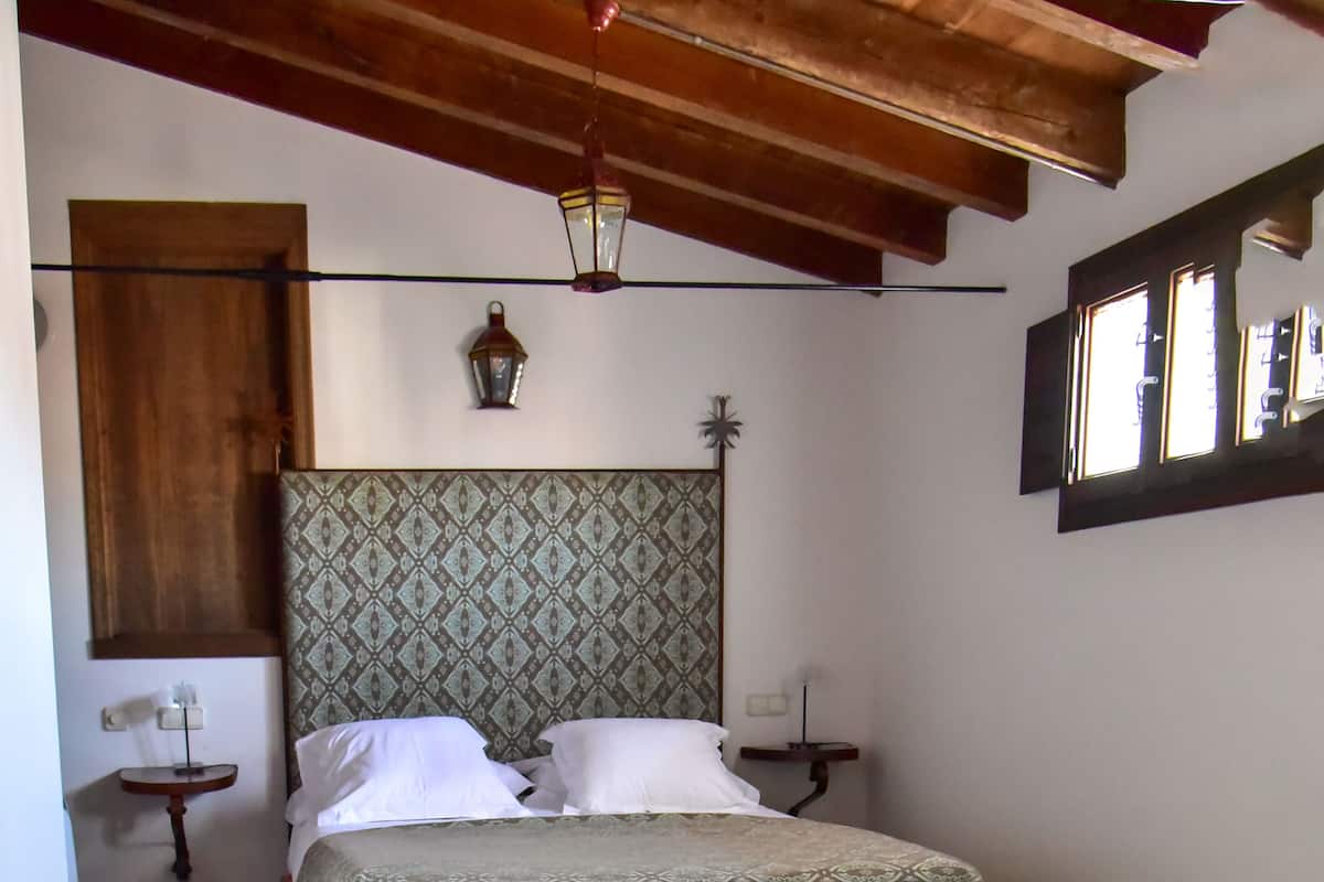 Un dormitorio con techo de madera, colcha estampada, mesita de noche con lámpara y ventana con persianas.