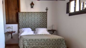 A bedroom with a wooden ceiling, a patterned bedspread, a bedside table with a lamp, and a window with blinds.