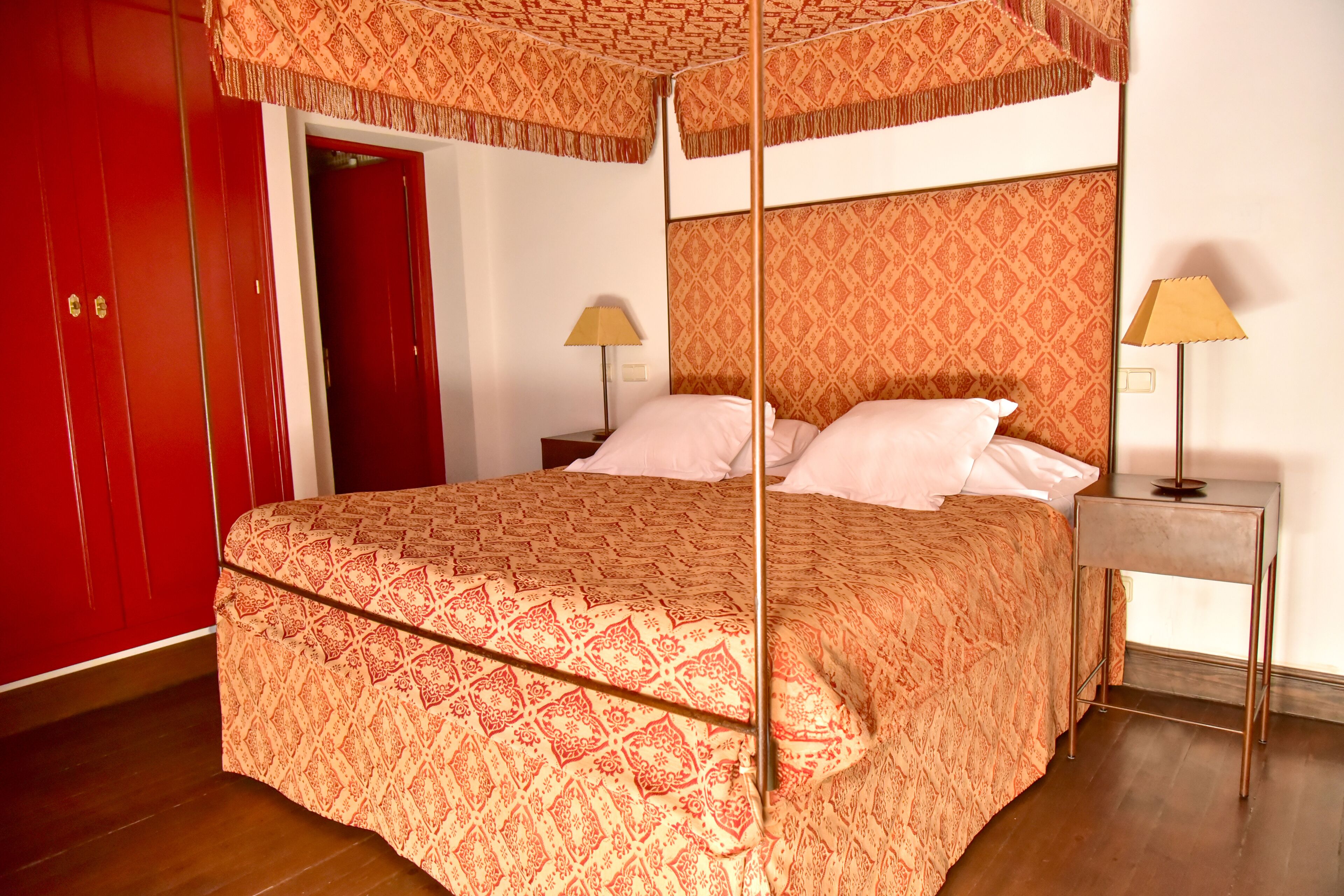 A four-poster bed with a patterned bedspread, two bedside tables with lamps, and a red wardrobe.