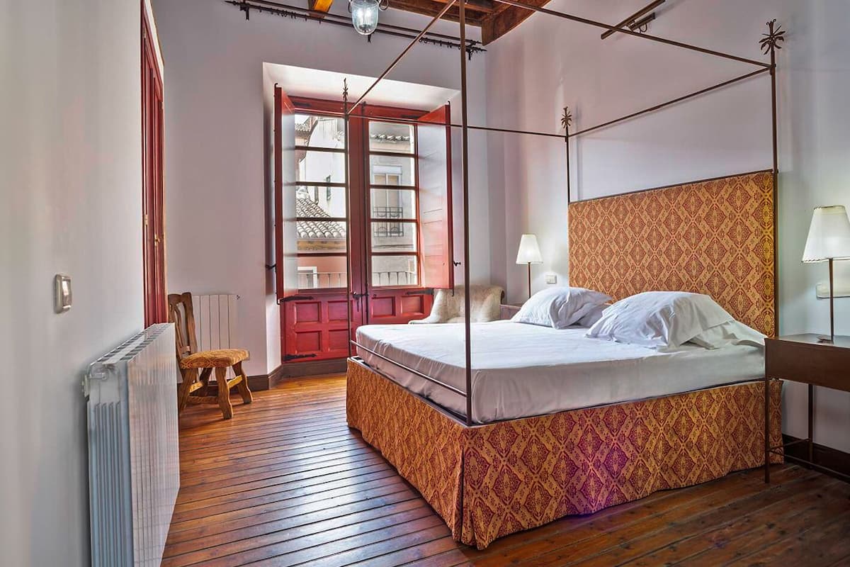 Una cama con dosel y un cabecero estampado, suelo de madera, un radiador y una puerta roja con paneles de vidrio.