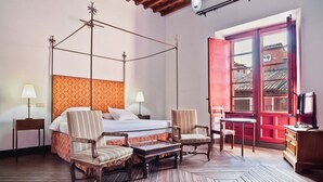 A hotel room with a four-poster bed, two chairs, a small table, a TV, and a large window with a view of buildings. - Palacio Rejadorada (Toro)