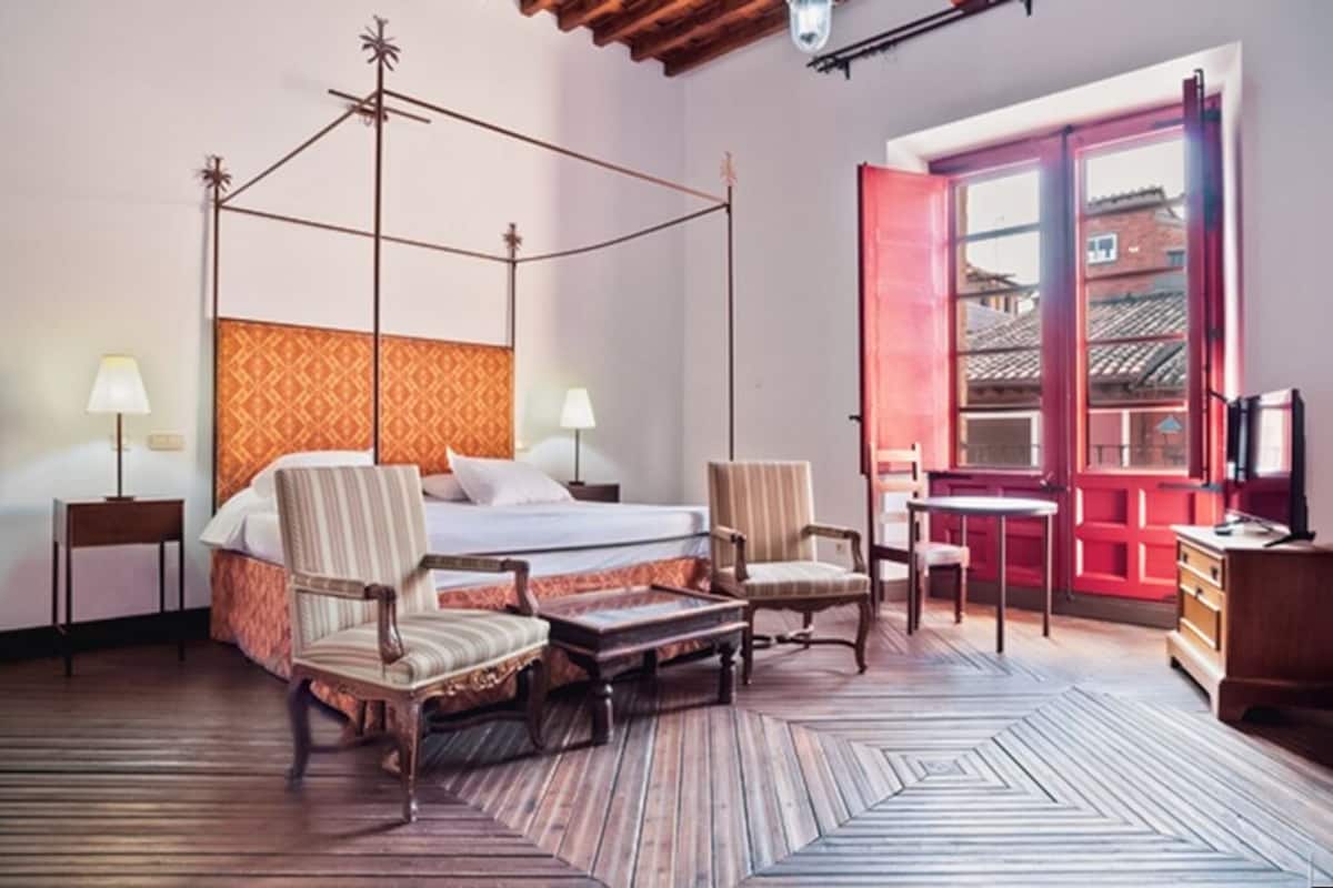 Habitación de hotel con cama con dosel, dos sillones, una mesita, televisor y un amplio ventanal con vistas a edificios.