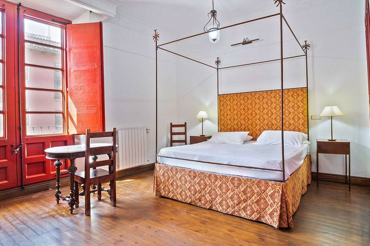 Una cama con dosel y tablero de cabecera estampado, una silla de madera, una mesita y una puerta roja.