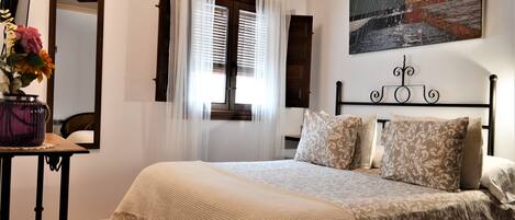 Un dormitorio con cama, una mesita de noche con un florero, una ventana con cortinas y un cuadro en la pared.