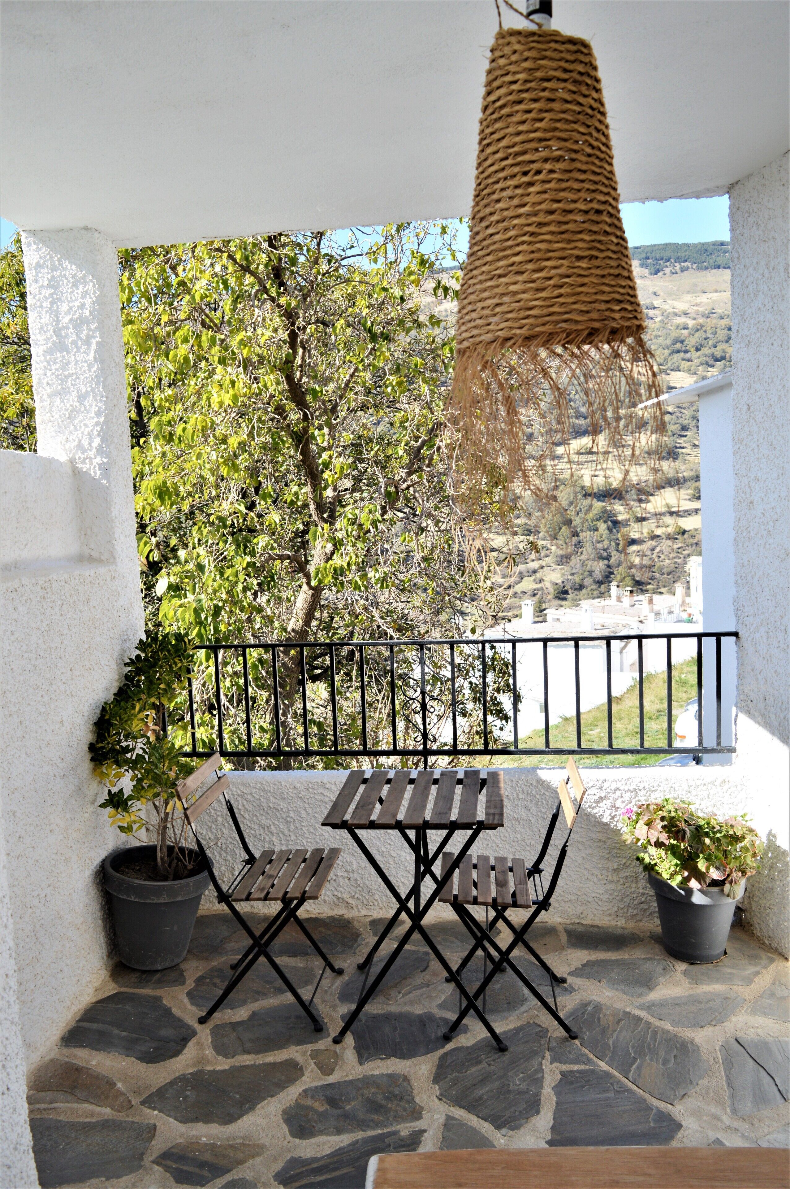 A balcony with a stone floor, a wooden table and chairs, a hanging light fixture, and potted plants.
