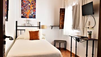Un dormitorio con cama, mesita de noche con lámpara, televisor montado en la pared y ventana con cortinas.