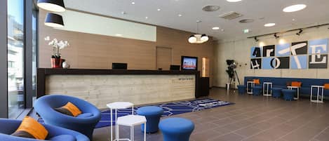 A modern hotel lobby with a reception desk, blue seating area, and a large window.
