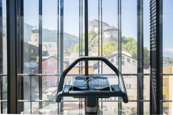 Una bicicleta estática con pantalla digital, ubicada frente a una ventana con vistas a edificios y una montaña.