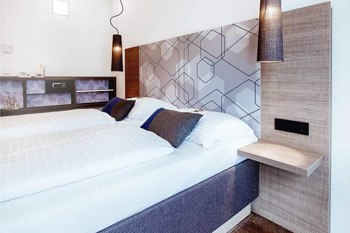 Ein modernes Hotelzimmer mit einem großen Bett, einem Kopfteil mit geometrischen Mustern, einem Nachttisch und einer hängenden Leuchte.