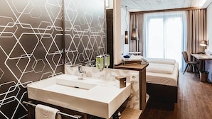 A modern hotel room with a large mirror, a white sink, a bed, a desk, and a window with curtains. - arte Hotel Kufstein (Kufstein)