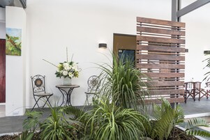 Un jardín interior moderno con un separador de madera, sillas de metal, una mesa con un jarrón de flores y diversas plantas.