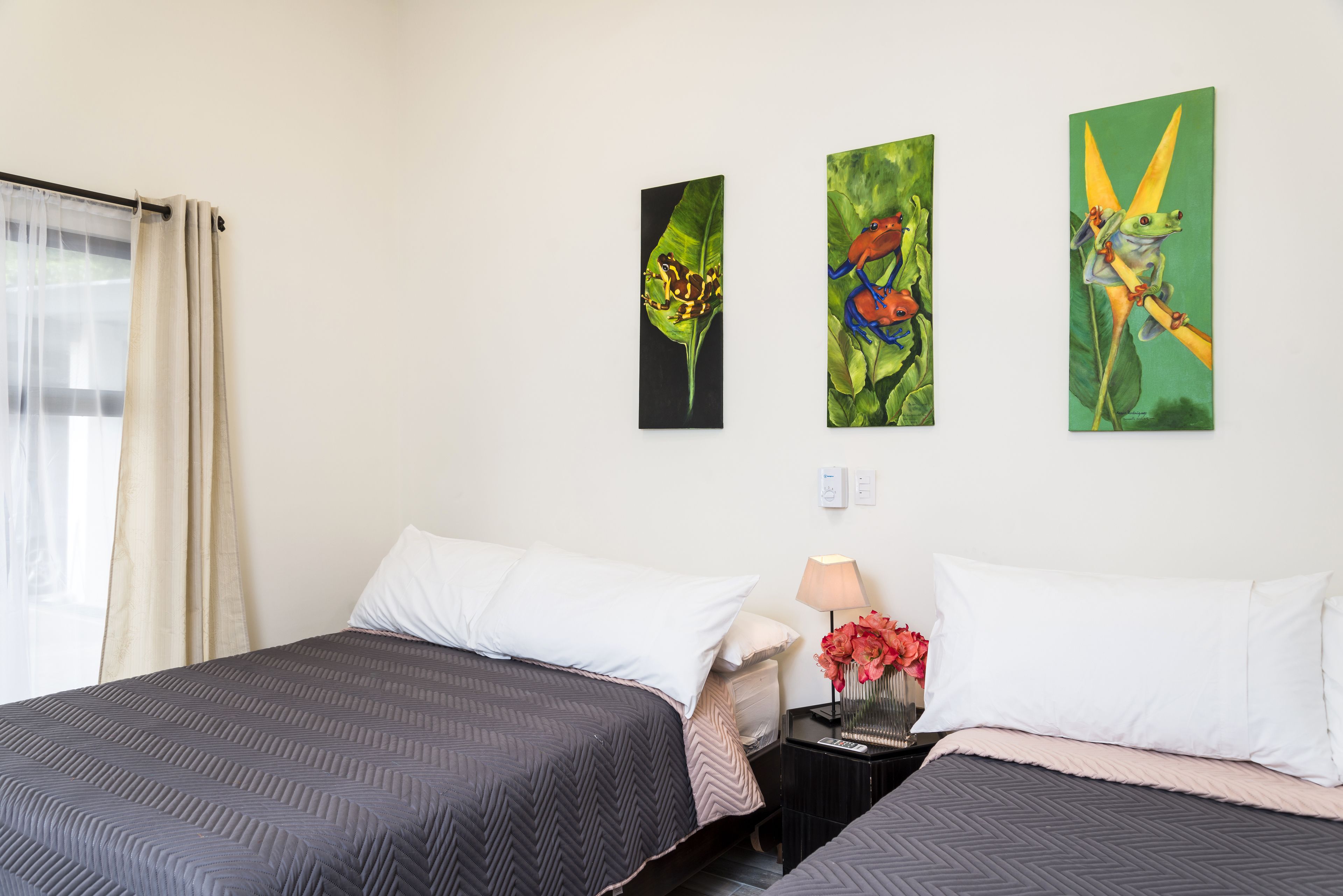 A hotel room with two beds, a nightstand with a lamp and vase, and three paintings on the wall.