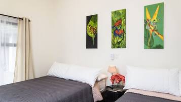 A hotel room with two beds, a nightstand with a lamp and vase, and three paintings on the wall.