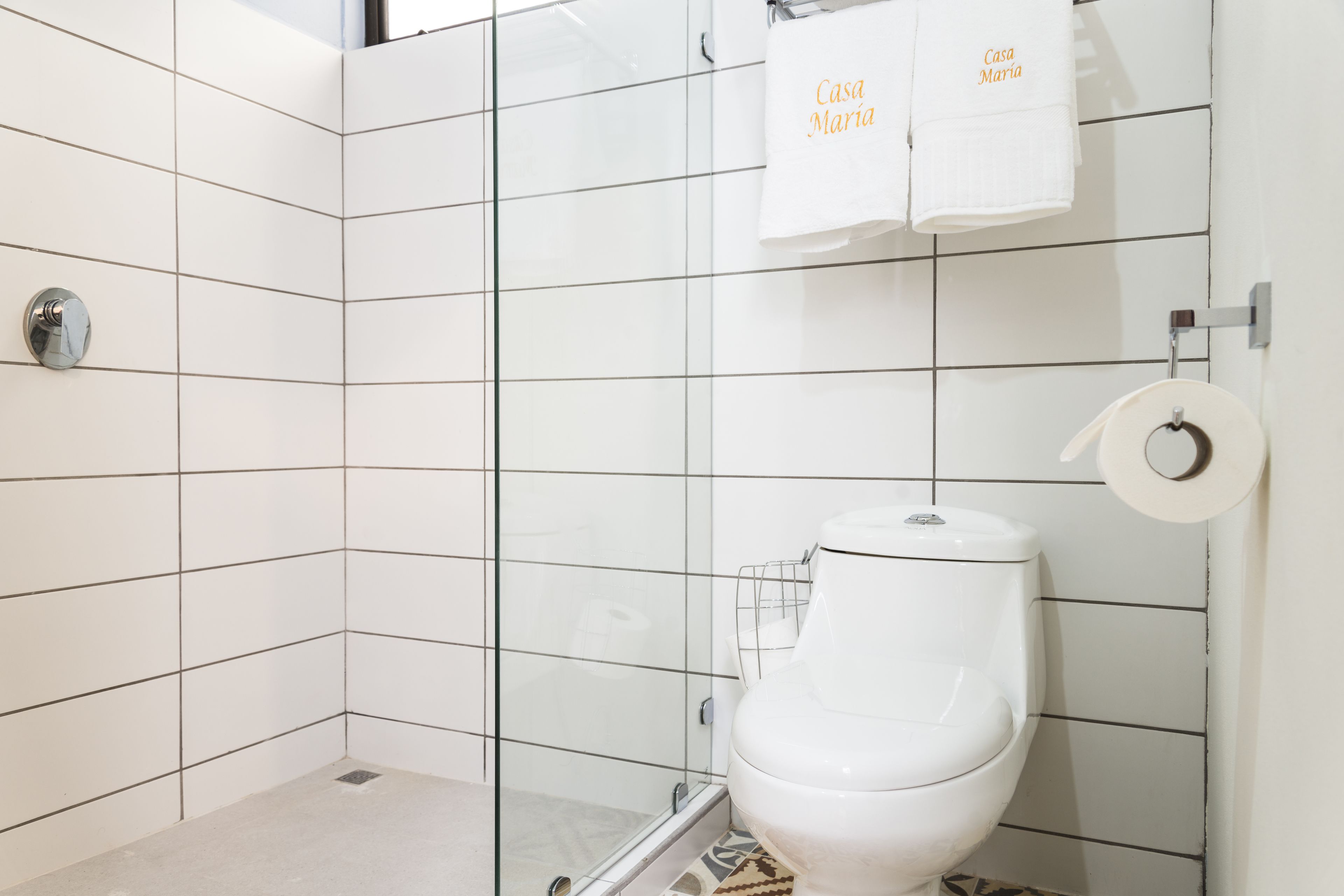 Un baño moderno con azulejos blancos, cabina de ducha de vidrio, inodoro y dos toallas colgadas en un perchero.