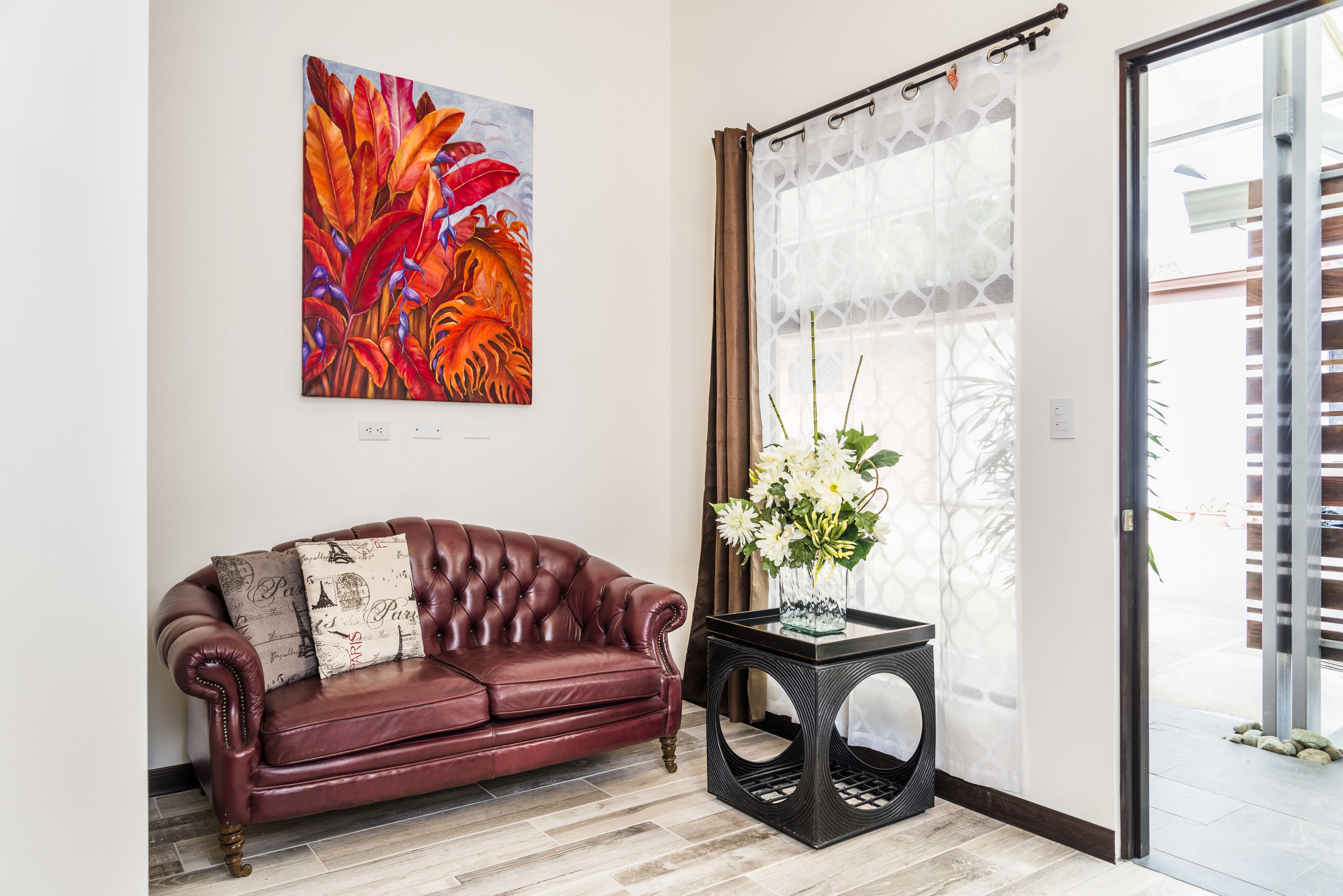 Un sofá de cuero rojo con cojines estampados, una mesita auxiliar negra con un jarrón de flores y un cuadro abstracto colorido en la pared.