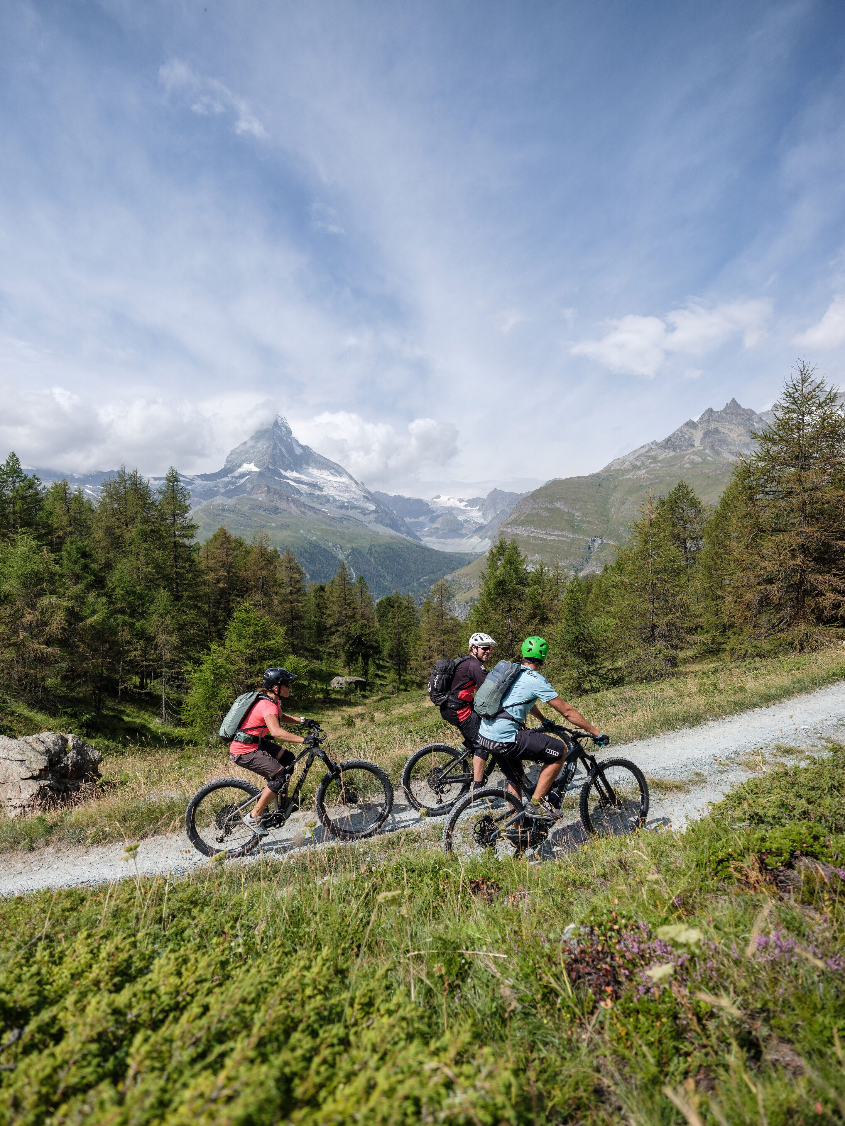 Three cyclists on a mountain trail with a view of snow-capped peaks in the background.