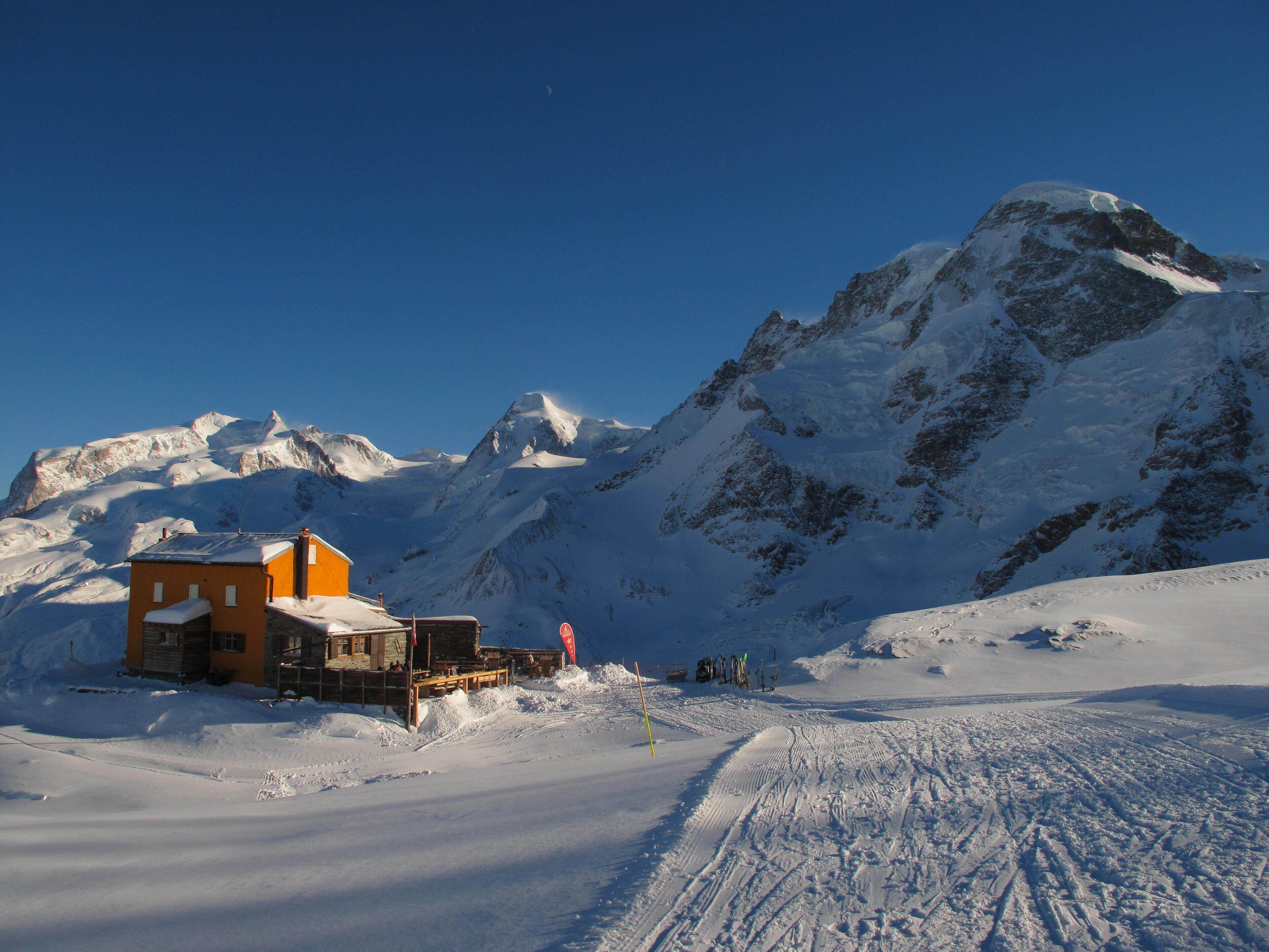 A snow-covered mountain with a small lodge featuring a wooden deck and a ski lift in the foreground.