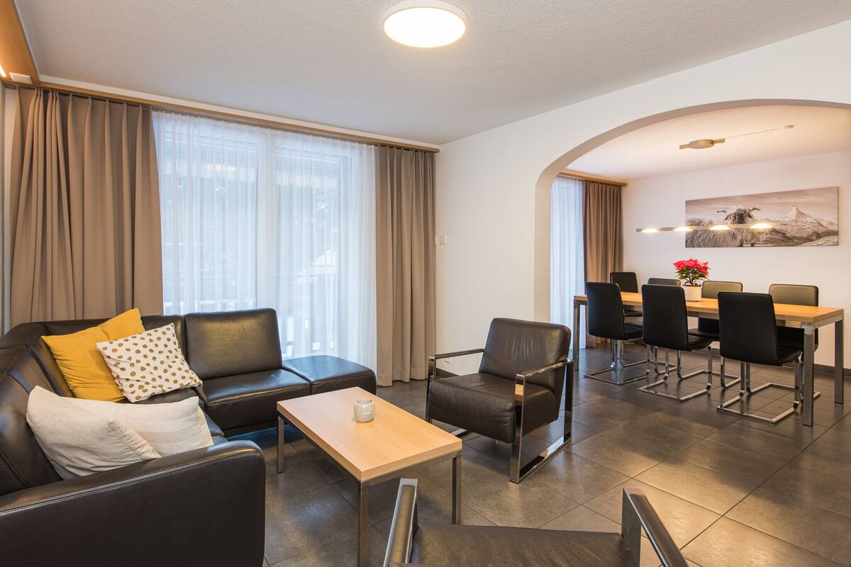 En moderne stue med sofa, lænestol og et lille bord. Der er et spiseområde med bord og stole.