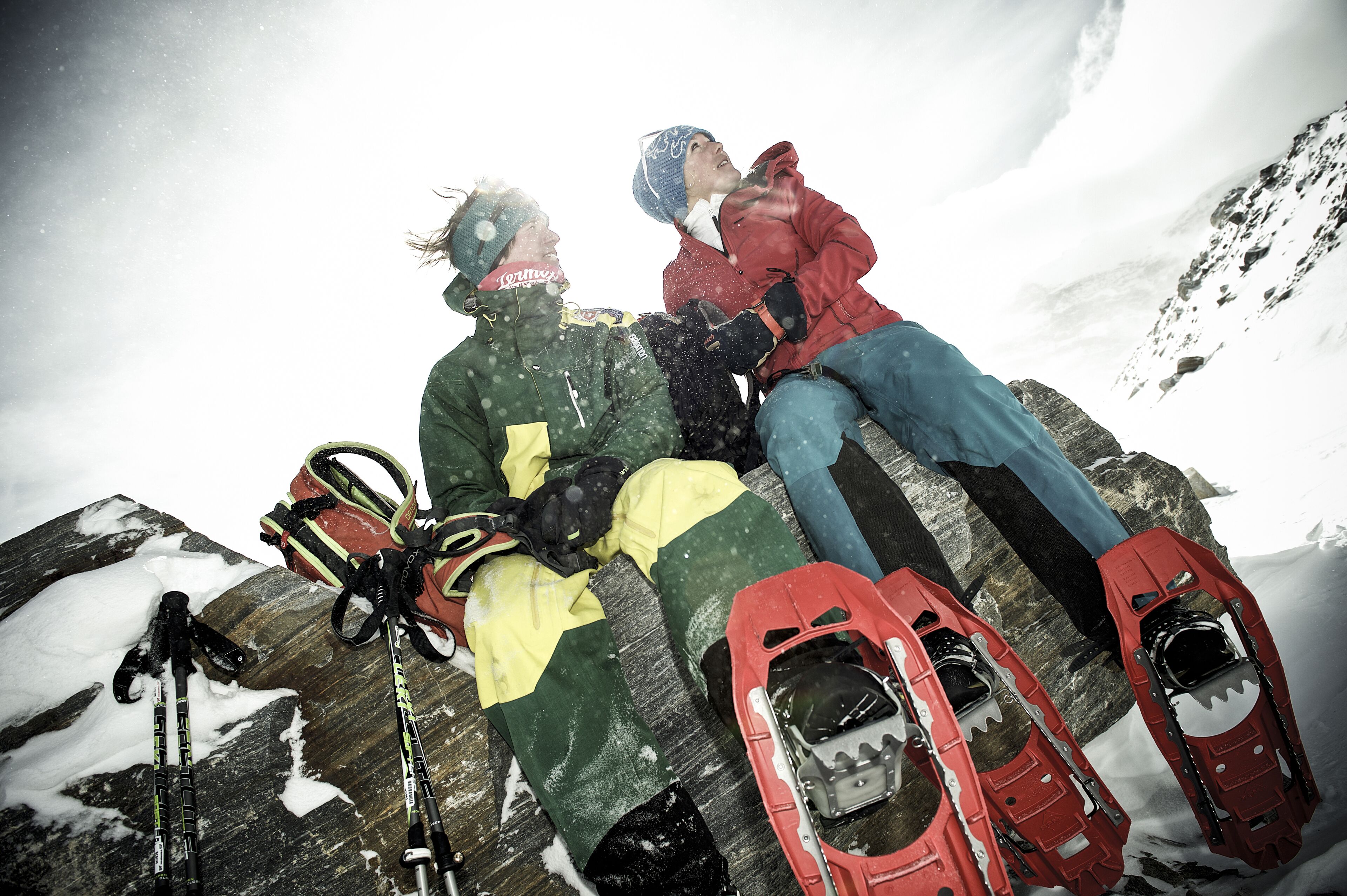 Two people sitting on a snowy mountain with snowshoes and ski poles around them.