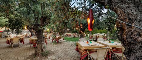 Un espace de restauration en plein air, avec des tables et des chaises placées sous les arbres, le tout entouré de verdure.