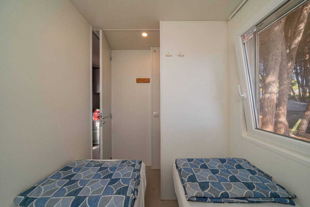 A small, narrow room with two beds, a window with a view of trees, and a door leading to a closet.