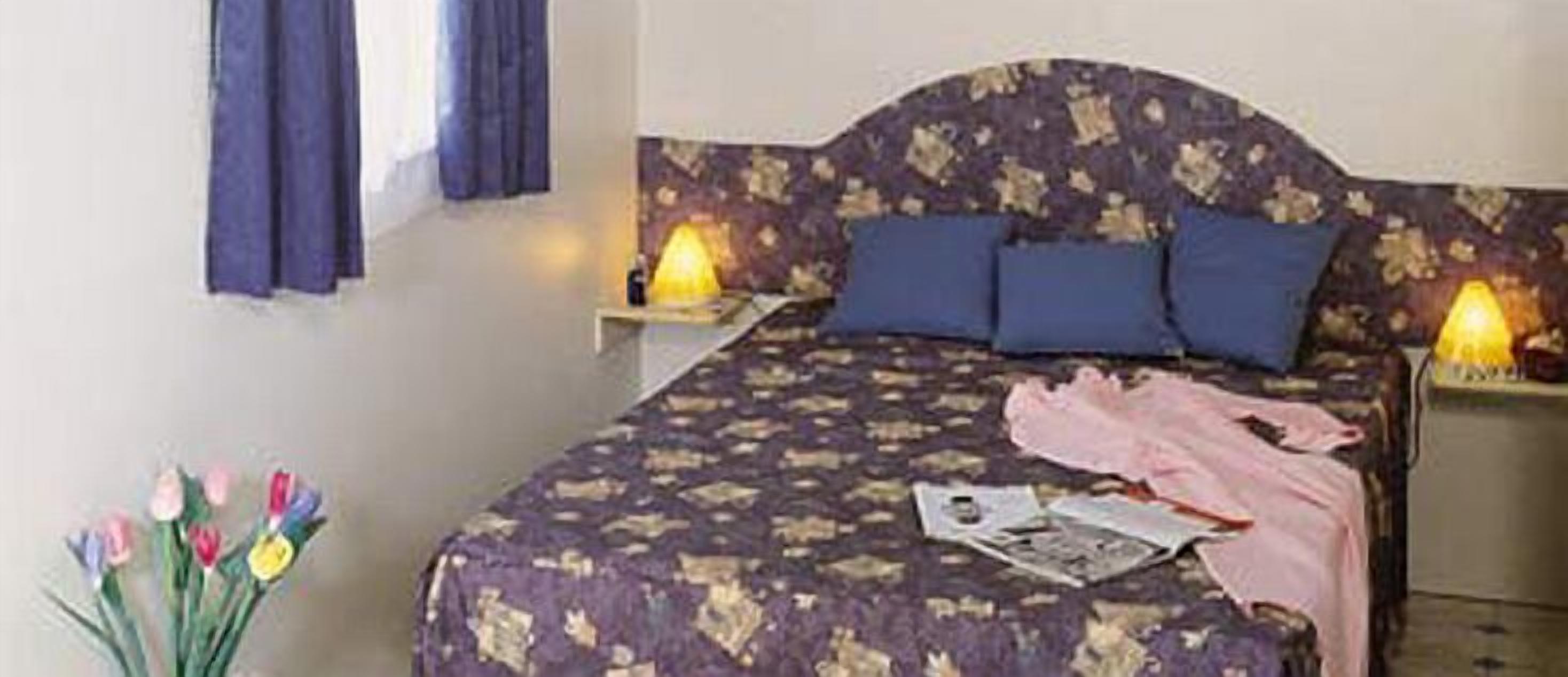 A bedroom with a floral bedspread, a bedside table with a lamp, and a window with curtains.