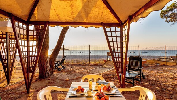 A beachside dining area with a table set for two, surrounded by chairs, under a canopy.