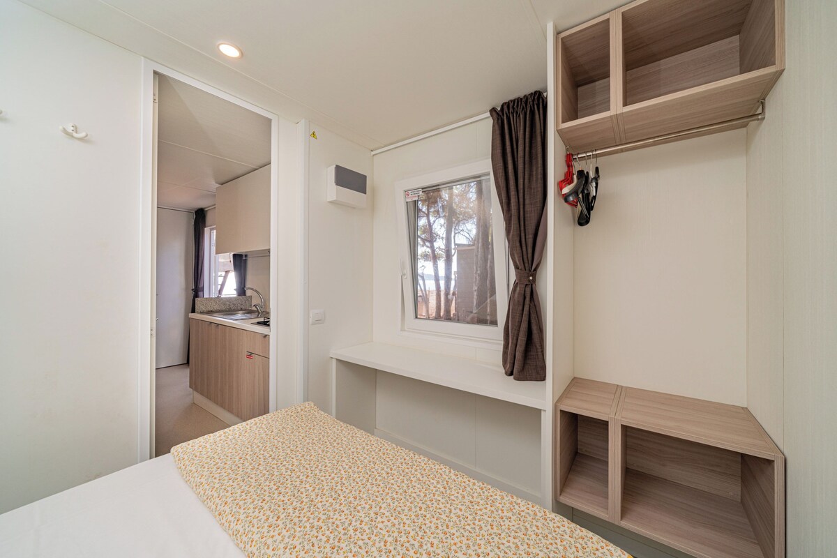 Un dormitorio con cama, ventana con cortinas, estante empotrado y vistas a la cocina y al baño.