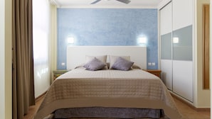 Une chambre à coucher comprenant un lit, deux tables de chevet, un ventilateur de plafond et une grande fenêtre avec des rideaux.