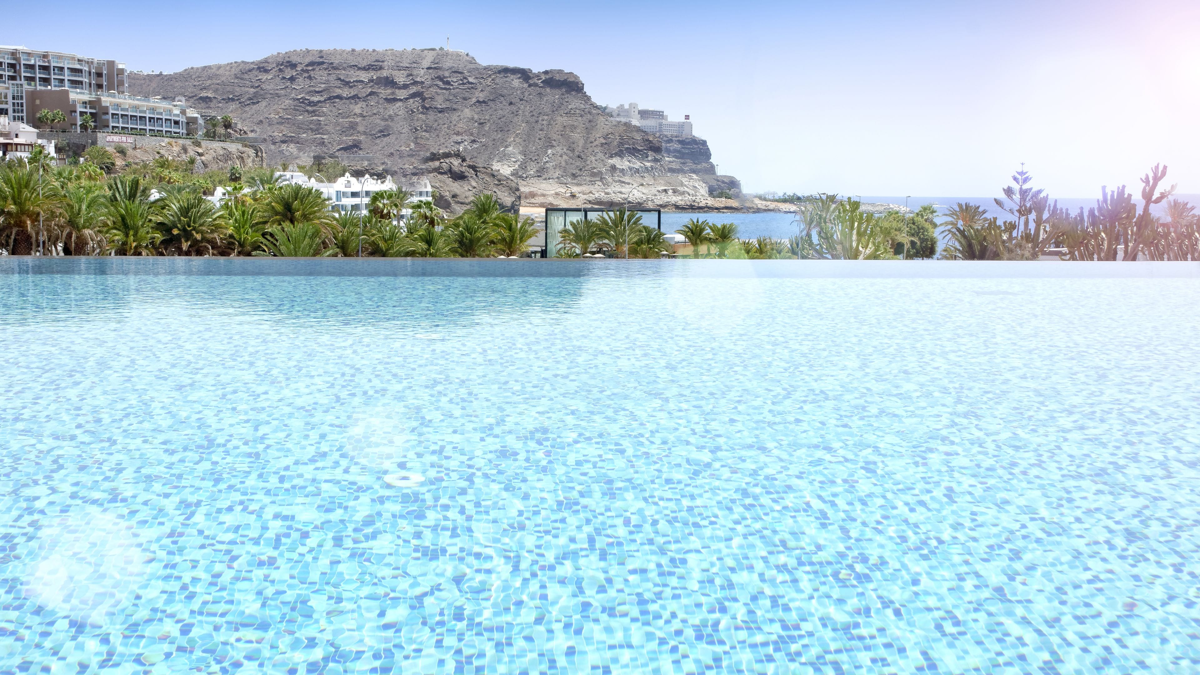 Una piscina azul cristalina con vistas a una montaña y edificios al fondo.
