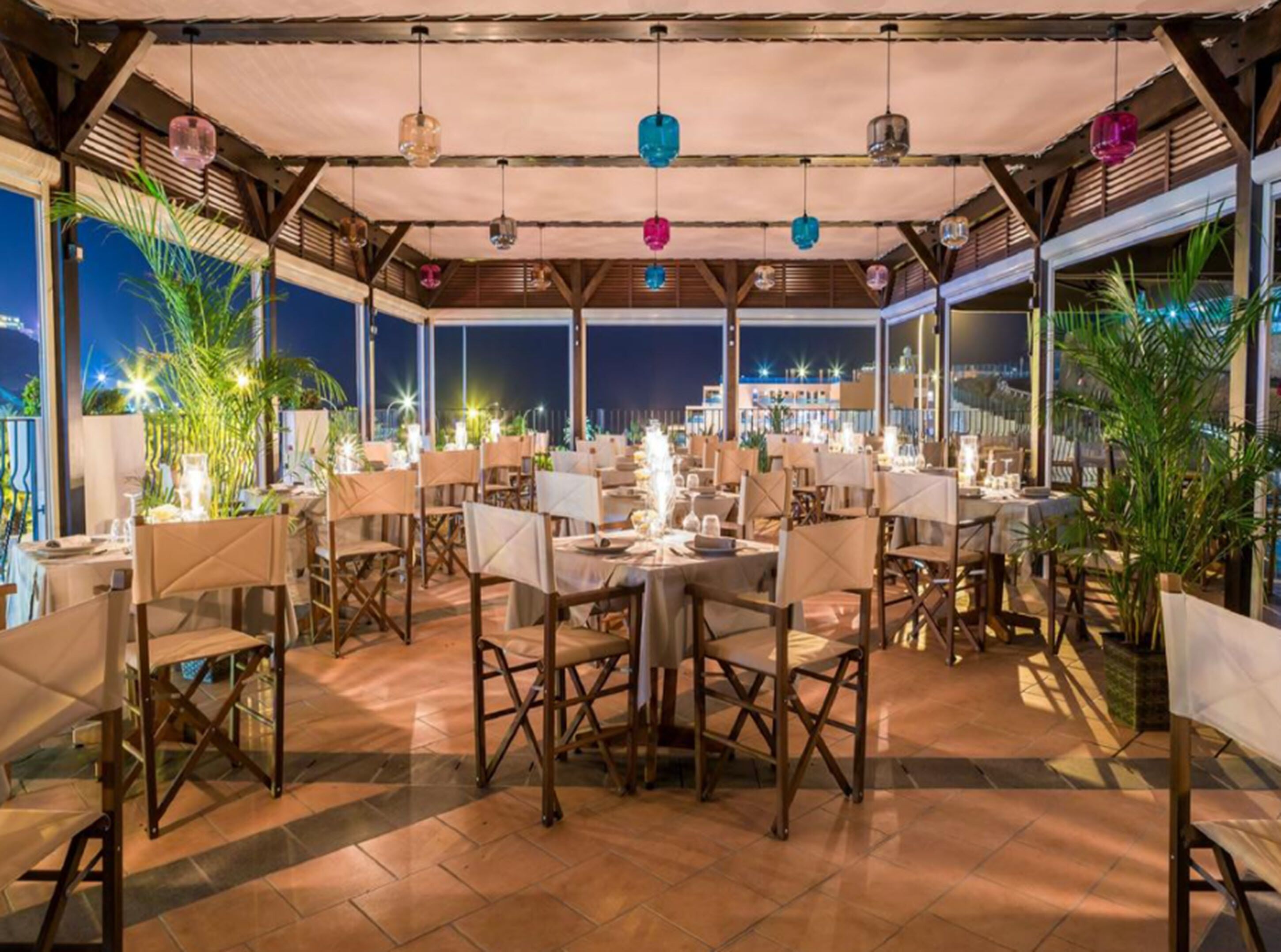 An outdoor dining area with tables and chairs, under a covered roof, with a view of the city at night.