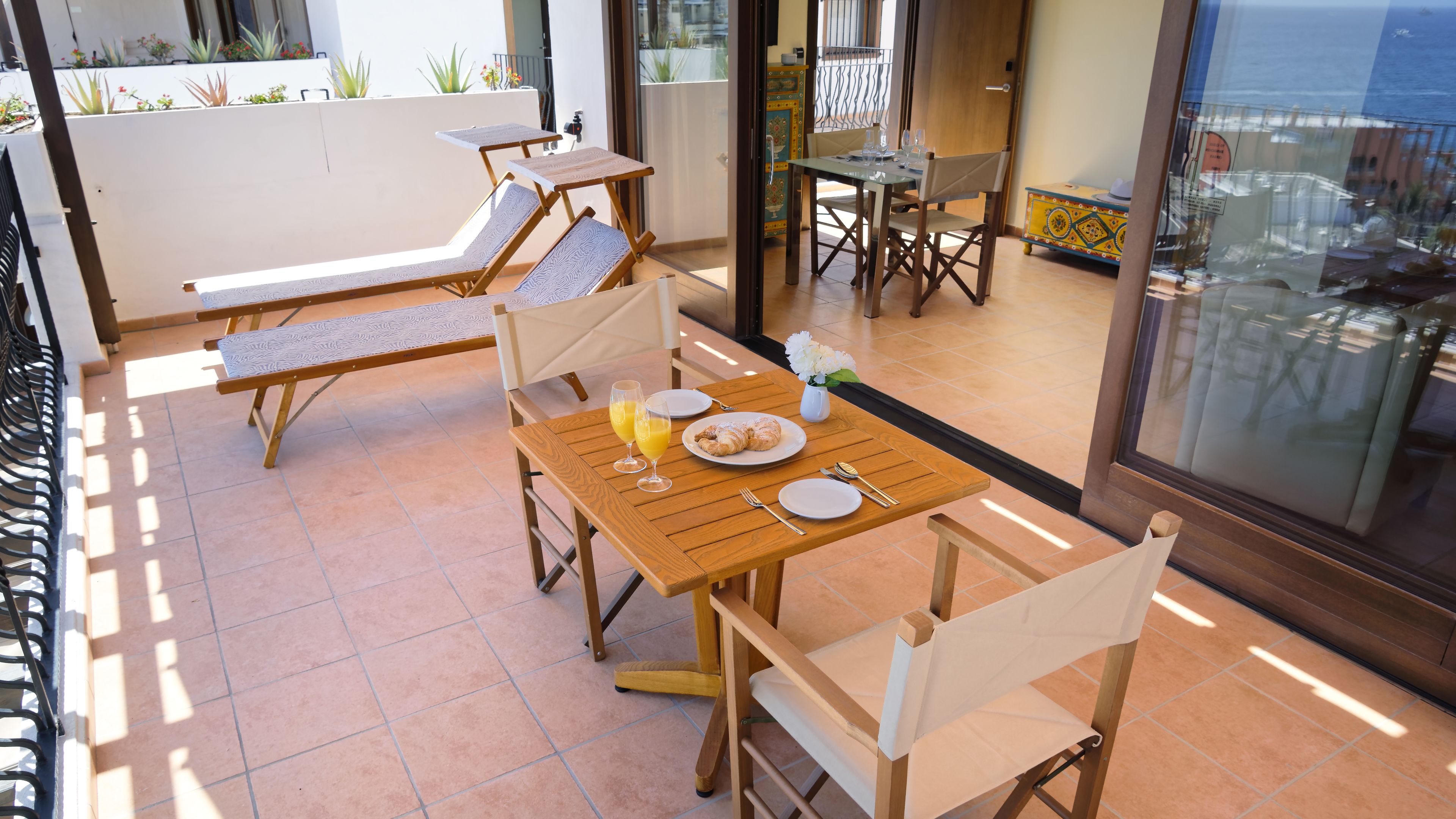 Un patio iluminado por el sol con muebles de madera, incluyendo una mesa lista para el desayuno con cruasanes y jugo de naranja.