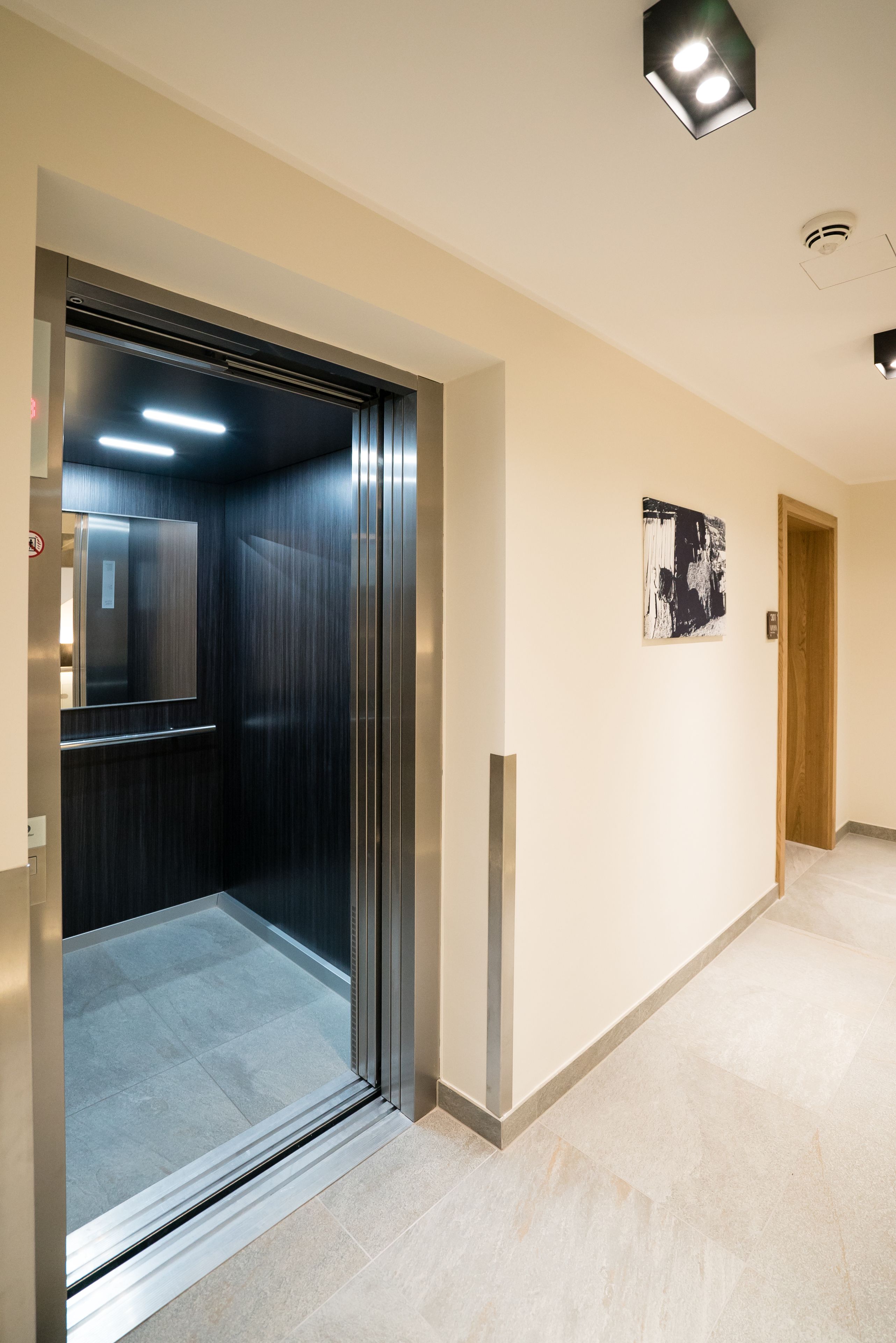 An elevator with a metallic interior and a glass door, leading to a hallway with light-colored walls and a tiled floor.