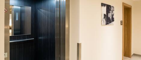 An elevator with a metallic interior and a glass door, leading to a hallway with light-colored walls and a tiled floor.